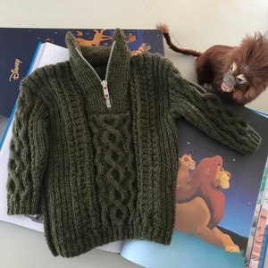 Op de afbeelding: Een groene gebreide trui met een rits en kabelbreipatroon.