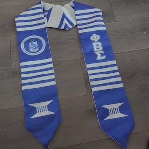 Könnte beinhalten: Ein blau-weißes Abschlussband mit horizontalen Streifen. Eine Seite zeigt ein rundes Emblem mit Text, die andere griechische Buchstaben. Das Band hat einen spitz zulaufenden Boden mit einer weißen Grafik.