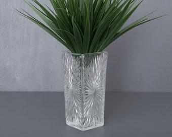 May include: A clear glass vase with a geometric pattern, filled with a green artificial grass plant.