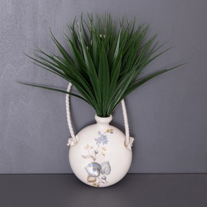 Puede incluir: Un jarrón de cerámica blanco con un diseño floral y un asa de cuerda. El jarrón está lleno de plantas artificiales verdes.