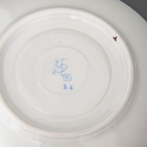 May include: Close-up of the bottom of a white porcelain saucer with a blue mark that reads "2-C 5" and a small blue square with the number "10" inside. The mark also includes the number "11" in blue.