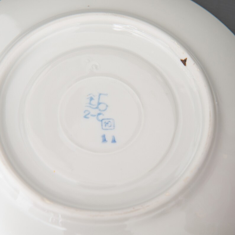 May include: Close-up of the bottom of a white porcelain saucer with a blue mark that reads "2-C 5" and a small blue square with the number "10" inside. The mark also includes the number "11" in blue.