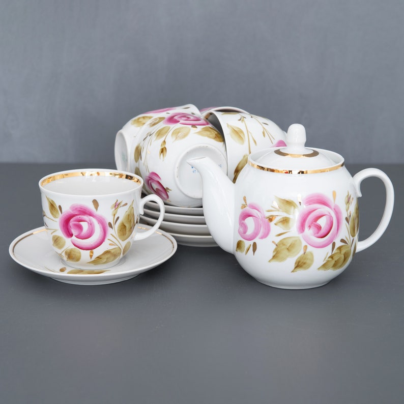 May include: A white porcelain tea set with a pink rose floral design and gold accents. The set includes a teapot, cups, and saucers.