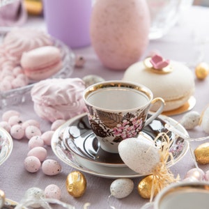 Könnte beinhalten: Eine weiße Teetasse mit Blumenmuster steht auf einer Untertasse mit braunem Rand. Die Teetasse ist umgeben von pastellrosa und weißen Marshmallows, gefleckten Ostereiern und einem weißen und goldenen Makron.