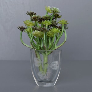 Puede incluir: Un jarrón de vidrio transparente con un diseño grabado contiene un ramo de suculentas artificiales. Las suculentas son una mezcla de tonos verdes y marrones.