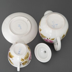 May include: A vintage tea set with a floral design. The set includes a teapot, a cup, a saucer, and a lid. The teapot and cup feature a pink rose design with green leaves. The saucer and lid are plain white.