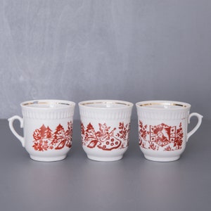 May include: Three white ceramic mugs with red illustrations and gold rims. Each mug features a different forest scene with figures and trees. The mugs have a classic design with a handle and a slightly ribbed texture.