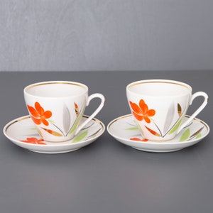 May include: Two white teacups and saucers with a floral design. The cups and saucers have a gold rim and feature orange flowers with green leaves on a white background.