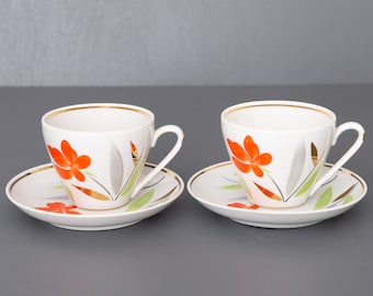 Puede incluir: Dos tazas de té blancas y platillos con un diseño floral. Las tazas y los platillos tienen un borde dorado y presentan flores naranjas con hojas verdes sobre un fondo blanco.