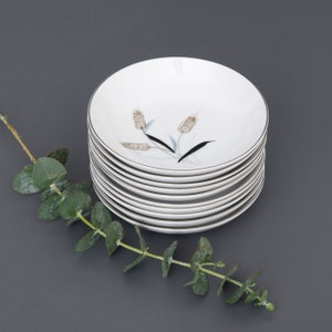 Könnte beinhalten: Ein Stapel weißer Keramik-Salatteller mit einem zarten Blumendesign mit braunen und schwarzen Weizenähren. Die Teller sind auf einer grauen Oberfläche gestapelt.