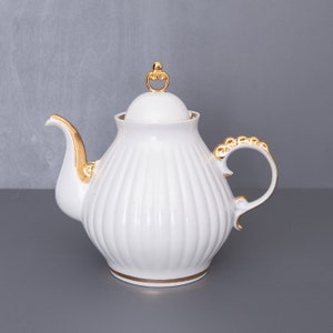 Op de afbeelding: Witte porseleinen theepot met een gouden deksel en handvat. De theepot heeft een geribbeld ontwerp en een gouden rand rond de basis.