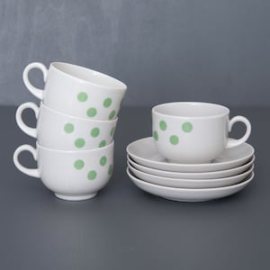 Puede incluir: Un juego de cuatro tazas de té blancas con lunares verdes, apiladas una encima de la otra. Una sola taza de té y un platillo con lunares verdes están sobre una superficie gris.