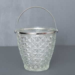 May include: Clear glass ice bucket with a geometric pattern and a silver metal rim and handle. The bucket has a flared shape and is designed to hold ice for chilling beverages. The handle arches over the top for easy carrying.