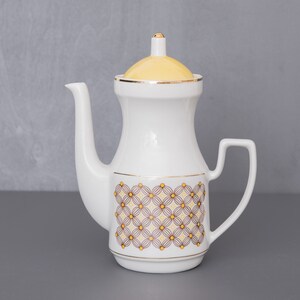 Könnte beinhalten: Weiße Keramik-Teekanne mit gelbem Deckel und einem geometrischen Muster in Braun und Gelb. Die Teekanne hat einen goldenen Rand um den Deckel und die Basis.