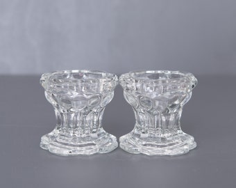 May include: Two clear glass candle holders with a textured, raised design. The holders have a wide base and a narrow top.