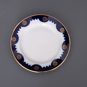 Puede incluir: Un platillo de porcelana blanca con un borde azul y dorado. El borde presenta un patrón repetitivo de flores estilizadas.