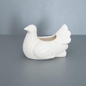 May include: A white ceramic dove-shaped planter. The bird has detailed feather textures and a small opening on its back for plants or flowers. The planter is a decorative item for home decor.