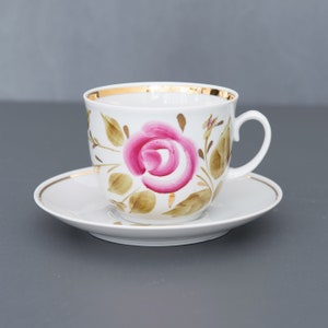 May include: A white porcelain teacup and saucer with a gold rim. The cup features a hand-painted pink rose with green leaves.