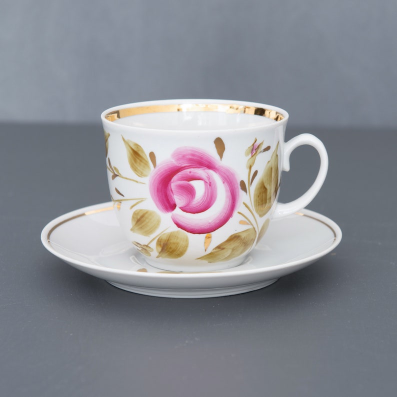 May include: A white porcelain teacup and saucer with a gold rim. The cup features a hand-painted pink rose with green leaves.