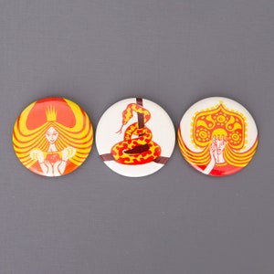 May include: Three round pinback buttons with colourful illustrations. The first button features a woman with a crown and a red heart. The second button features a yellow snake with red markings. The third button features a woman wearing a red and yellow headdress.