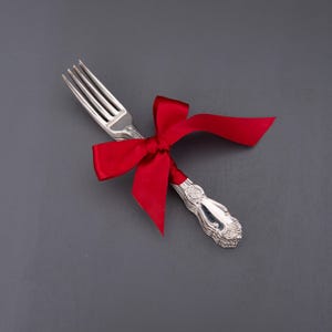Può includere: Una forchetta d'argento legata con un vivace nastro di raso rosso. La forchetta ha un manico decorativo ed è presentata su uno sfondo grigio scuro, suggerendo un regalo o un'occasione speciale.