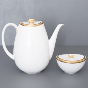 Puede incluir: Tetera y azucarero de porcelana blanca con ribetes dorados. La tetera tiene un cuerpo redondeado y un pico. El azucarero tiene una tapa con un pomo.