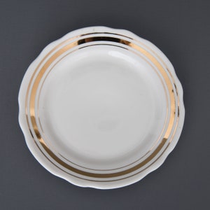 May include: A white ceramic plate with a scalloped edge and three gold bands around the rim.