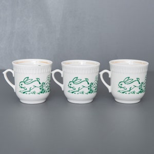 May include: Three white ceramic mugs with green bunny illustrations. Each mug has a handle and a green bunny running with a green leafy background.