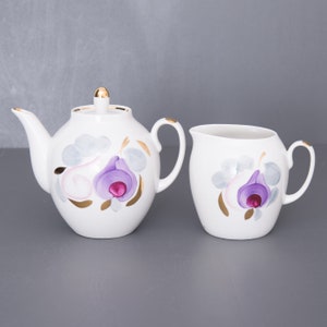 May include: White ceramic teapot and creamer set. The teapot has a lid with a gold finial and a curved spout. Both pieces are decorated with a purple and pink floral design with gold accents. The set is ideal for serving tea.