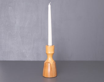 Puede incluir: Un portavelas de madera con una vela blanca. El portavelas es de color marrón claro y tiene un diseño sencillo.