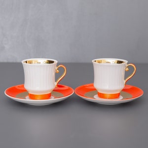 Könnte beinhalten: Zwei weiße Porzellan-Kaffeetassen mit goldenen Verzierungen und orangen Untertassen. Die Tassen haben ein geriffeltes Design und stehen auf einer grauen Oberfläche.