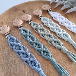 May include: Five macrame dummy clips in shades of blue, green, and white. Each clip features a wooden button and a metal clasp. The macrame cords are intricately woven, with a tassel at the end of each clip. The clips are arranged on a wooden surface.
