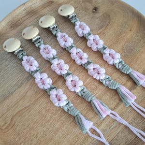 May include: Four baby pacifier clips with wooden ends, silver metal clips, and macrame detailing. The macrame is light green with pink flower accents and pink tassels. The clips are arranged on a wooden surface.