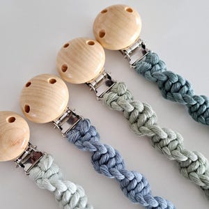 May include: Four pacifier clips with wooden beads and braided cords in shades of blue and green. Each clip has a silver-coloured metal clasp. The wooden beads have three holes.
