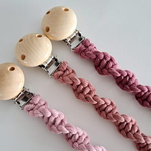 May include: Three pacifier clips. Each has a wooden round top and a braided cord in shades of pink and mauve. The clips have silver metal clasps. The wooden tops have three holes.