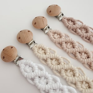 May include: Four macrame dummy clips in white, cream, and tan. Each clip features a wooden round top and a metal clip. The straps are made of woven cotton, with a visible macrame design.