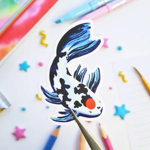 May include: A sticker featuring a koi fish design. The fish has a white body with black spots and a blue and black tail. It has a red eye and is being held by tweezers. The background includes colorful pencils and star-shaped decorations.