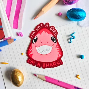 Könnte beinhalten: Ein pinkfarbener Hai-Aufkleber mit dem Text "I'M NOT A GIRL I'M A SHARK!" wird auf einer linierten Papieroberfläche angezeigt. Der Aufkleber hat ein lächelndes Haigesicht mit weißen Zähnen und schwarzen Augen. Stifte und bunte Dekorationsartikel umgeben den Aufkleber.