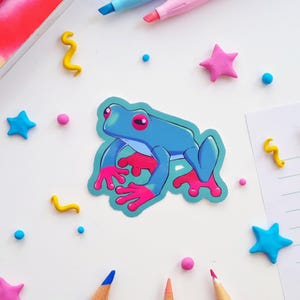 May include: A blue sticker of a cartoon frog with pink feet and red eyes. The sticker is surrounded by colorful star and dot-shaped decorations, along with pencils and markers, all on a white surface.