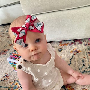Peut inclure: Un bébé portant une combinaison en tricot blanc avec des pompons blancs et un bandeau rose avec un nœud représentant le contour d'un état et le mot "Ohio".