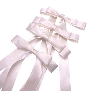 May include: Four white satin ribbon bows with long tails. The bows are arranged in a stack, with the largest bow at the bottom and the smallest bow at the top.