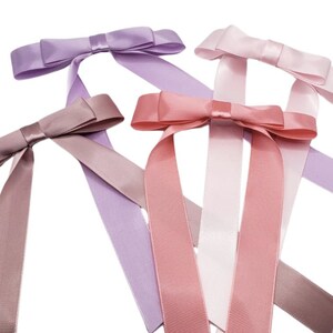 May include: Four satin ribbon bows in different shades of pink and purple. The bows are tied with long tails and are arranged in a criss-cross pattern.