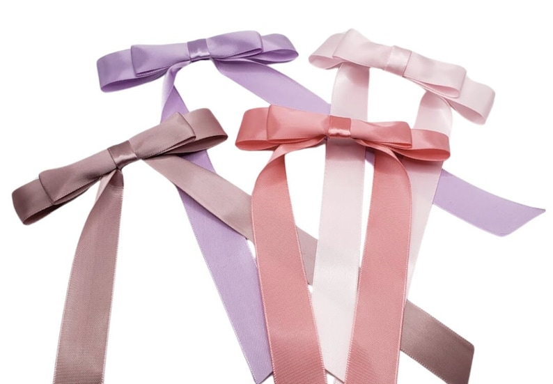 May include: Four satin ribbon bows in different shades of pink and purple. The bows are tied with long tails and are arranged in a criss-cross pattern.