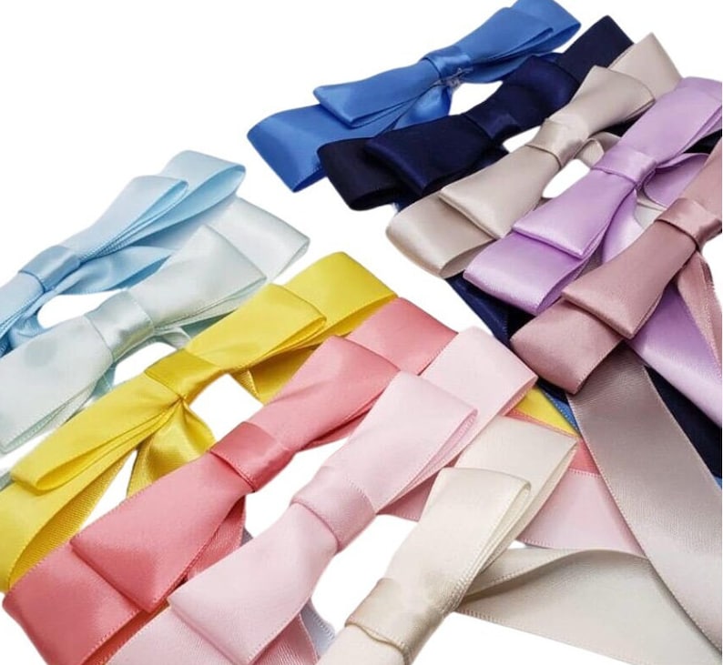 May include: Assortment of satin ribbon bows in various colours, including light blue, yellow, pink, and white. The bows are arranged in a random pattern on a white background.