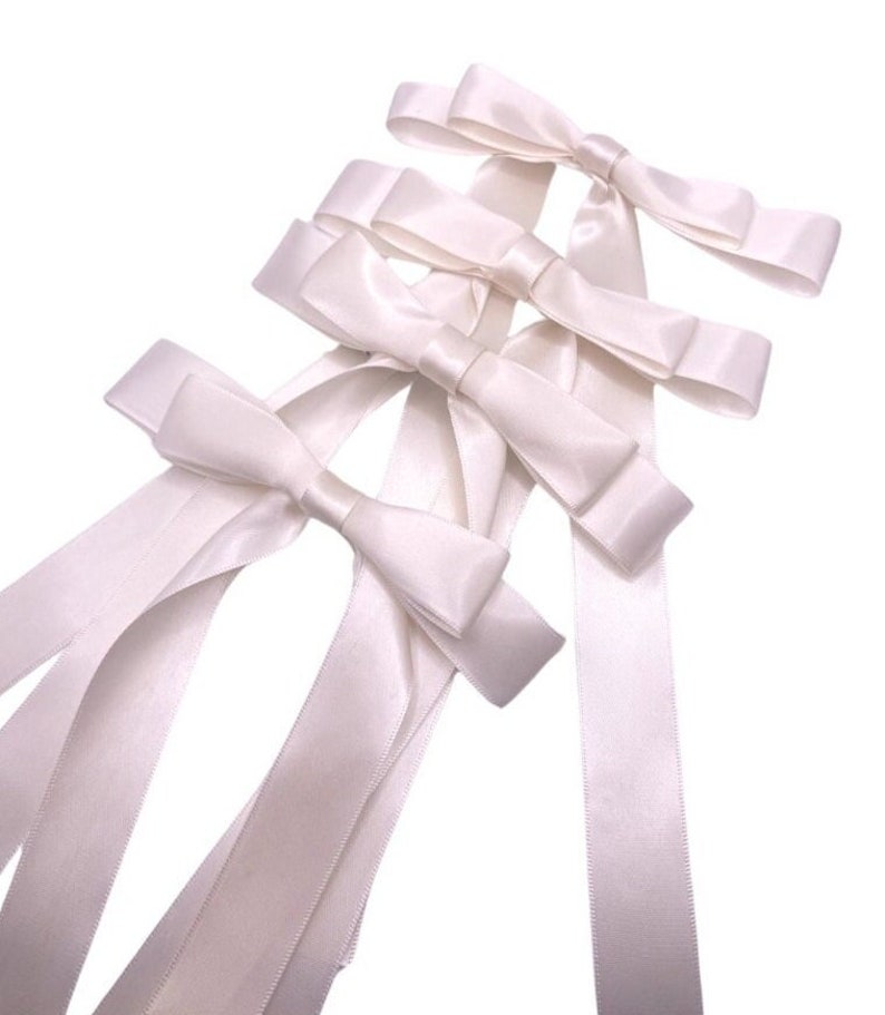 May include: Four white satin ribbon bows with long tails. The bows are arranged in a stack, with the largest bow at the bottom and the smallest bow at the top.