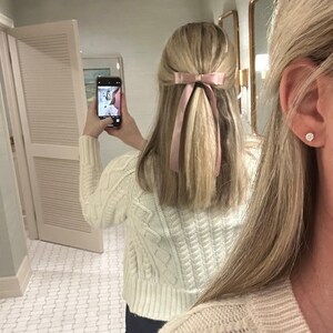 May include: A woman with long blonde hair wearing a white cable knit jumper and a pink ribbon in her hair. She is standing in a bathroom with white tiled floors.