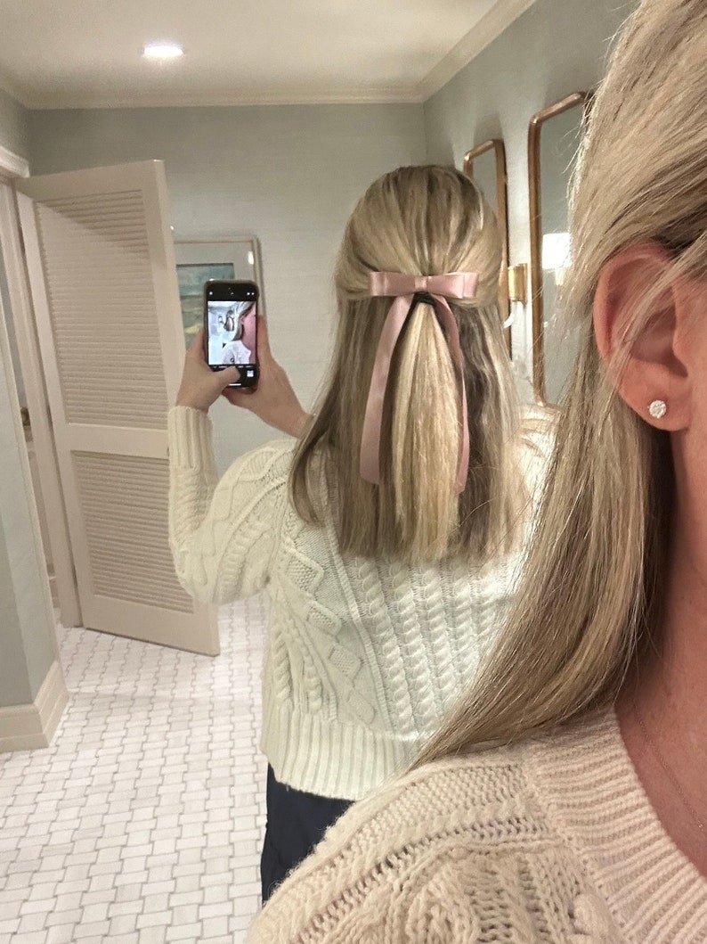 May include: A woman with long blonde hair wearing a white cable knit jumper and a pink ribbon in her hair. She is standing in a bathroom with white tiled floors.