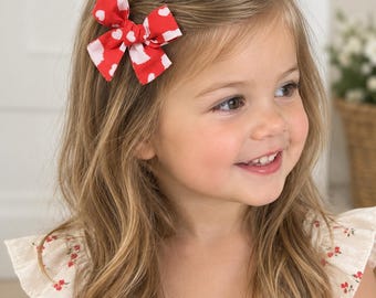 May include: A fabric hair bow featuring a vibrant red background with a repeating pattern of white hearts. The bow is constructed with a central knot detail and is secured to the hair, showcasing a classic shape with four distinct loops.