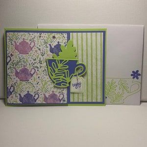 May include: A handmade greeting card with a purple teacup and green leaves on a white background with a green and white striped pattern. The card has a tag that says "enjoy".
