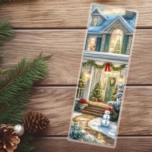 May include: A Christmas-themed bookmark featuring a detailed illustration of a snow-covered house with a decorated front door and a snowman. The house has a blue roof and green shutters, with a Christmas tree visible through the window.